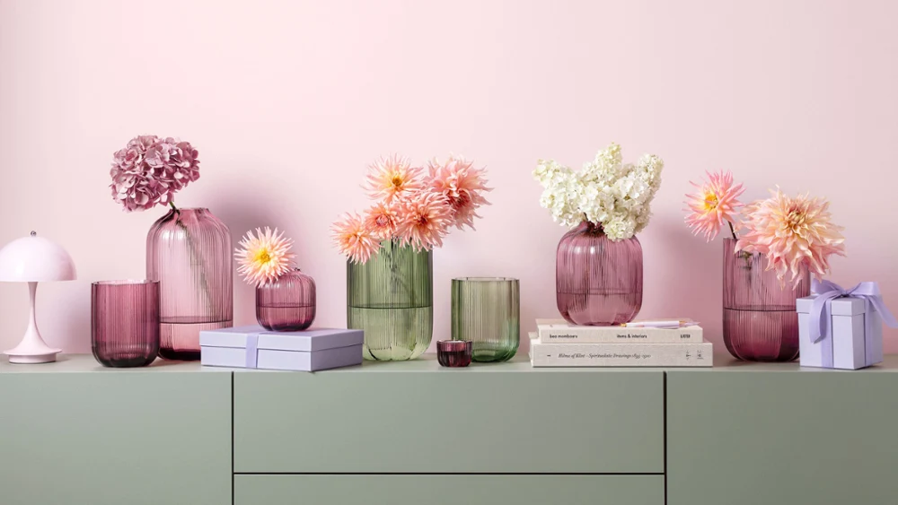 Various pastel-colored glass vases from Fleur stand on a green cabinet with stacked books and small gift boxes in front of a pink wall.