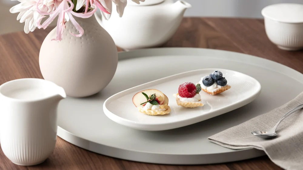 Een wit Afina bord met drie kleine gebakjes en fruit staat op een rond dienblad naast een bloemenvaas, een theepot en een melkkannetje op een houten tafel.
