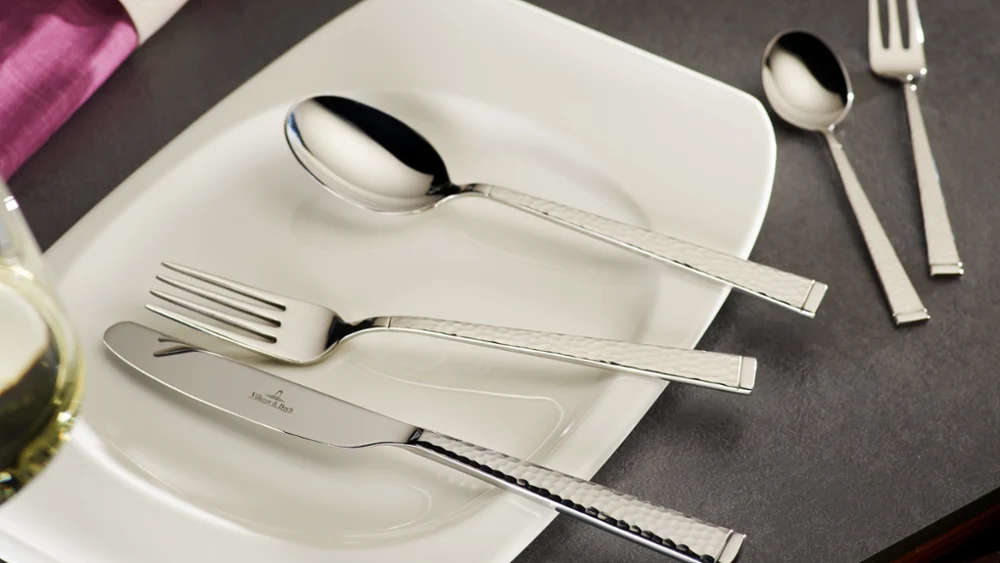 Op een wit rechthoekig bord van Villeroy & Boch Blacksmith liggen een glanzende vork, mes en lepel op een grijs oppervlak. Ernaast liggen een kleinere lepel en een vork uit de Blacksmith collectie. Een wijnglas en een roze servet zijn gedeeltelijk zichtbaar.