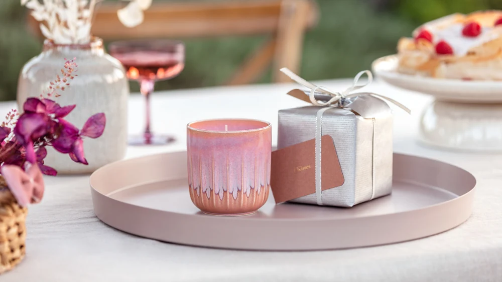 Geurkaars uit de collectie like. by Villeroy & Boch als cadeau op een rond dienblad met een zilveren cadeaubox op de achtergrond en roze orchideeën op de voorgrond.