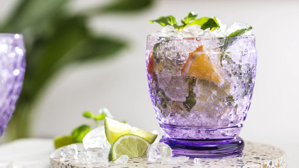 Ein Cocktail in einem violetten Boston Wasserglas, garniert mit Minze und Zitrusfrüchten, umgeben von Eiswürfeln und Limettenscheiben auf einem gesprenkelten Steintablett.
