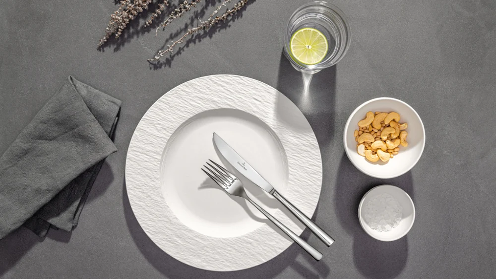 Een wit bord met vork en mes, een glas water met citroen, een schaaltje crackers en een servet op een grijze tafel.