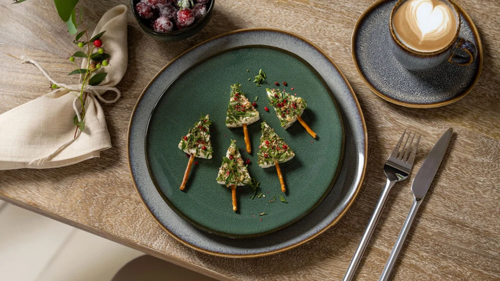 Platos de gres Lave de Villeroy & Boch con aperitivos de queso con forma de pino, capuchino y arándanos escarchados sobre mesa de madera.