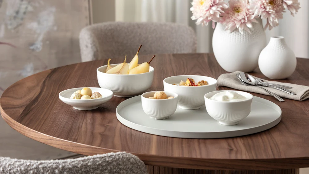 Een ronde houten tafel, gedekt met Afina-schalen met hapjes, een Manufacture-bloemenvaas en een gevouwen servet.