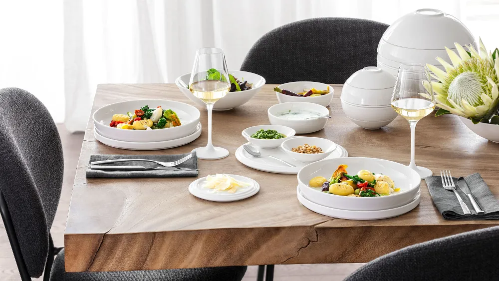 A table set with La Boule and black and gray chairs around it A table set with La Boule with two plates full of food, two glasses of white wine, various bowls with side dishes, and a flower arrangement with black and gray chairs around it.