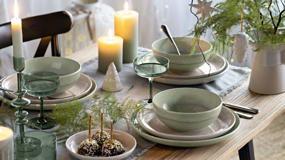 Una mesa navideña con platos, cuencos y vasos Perlemor de Villeroy & Boch en verde y beige, decorada con velas, una estrella y un pequeño árbol de Navidad. La mesa está decorada con un camino de mesa gris claro.