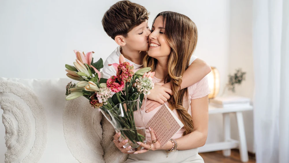 Een jongen omhelst zijn moeder. Ze houdt een vaas met bloemen en een ingepakt cadeau in haar handen