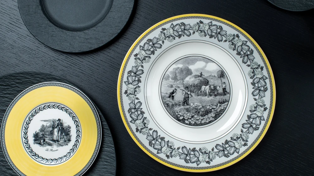 Audun and Manufacture Rock plates on a black table Elegant plates on a dark surface; one large with a rural scene and floral border, another smaller with a yellow rim depicting a couple.