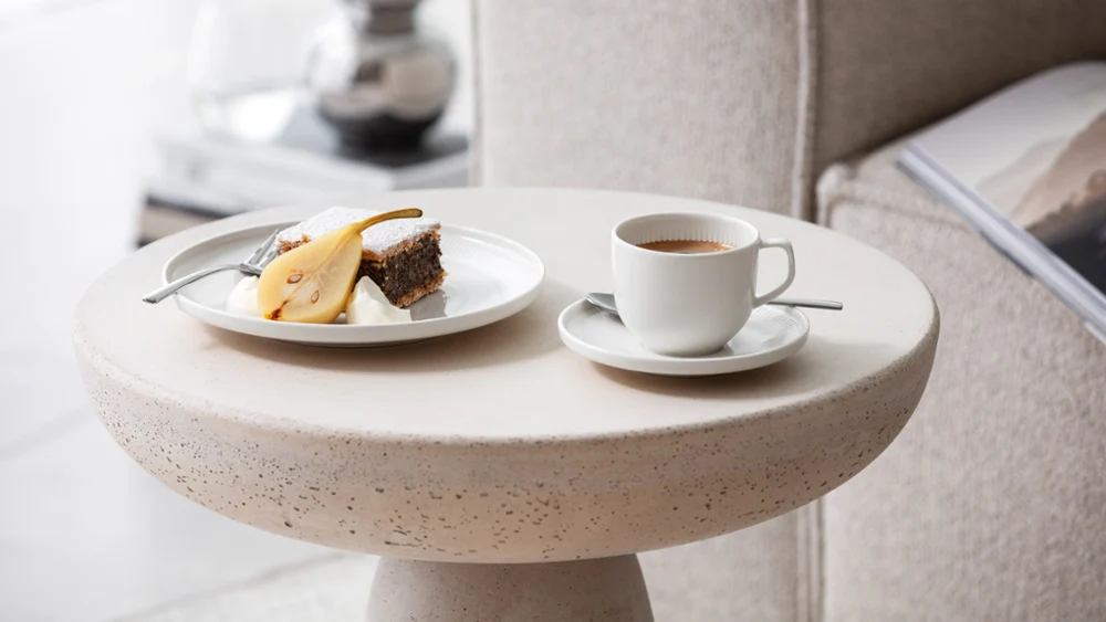 Een kopje koffie en een bordje cake geserveerd op een moderne bijzettafel met Afina servies van Villeroy & Boch.