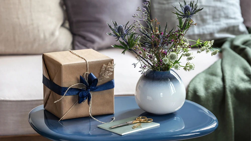 Su un tavolo blu c'è un regalo avvolto in carta marrone con un nastro blu, un vaso bianco di lavanda con fiori e un biglietto.
