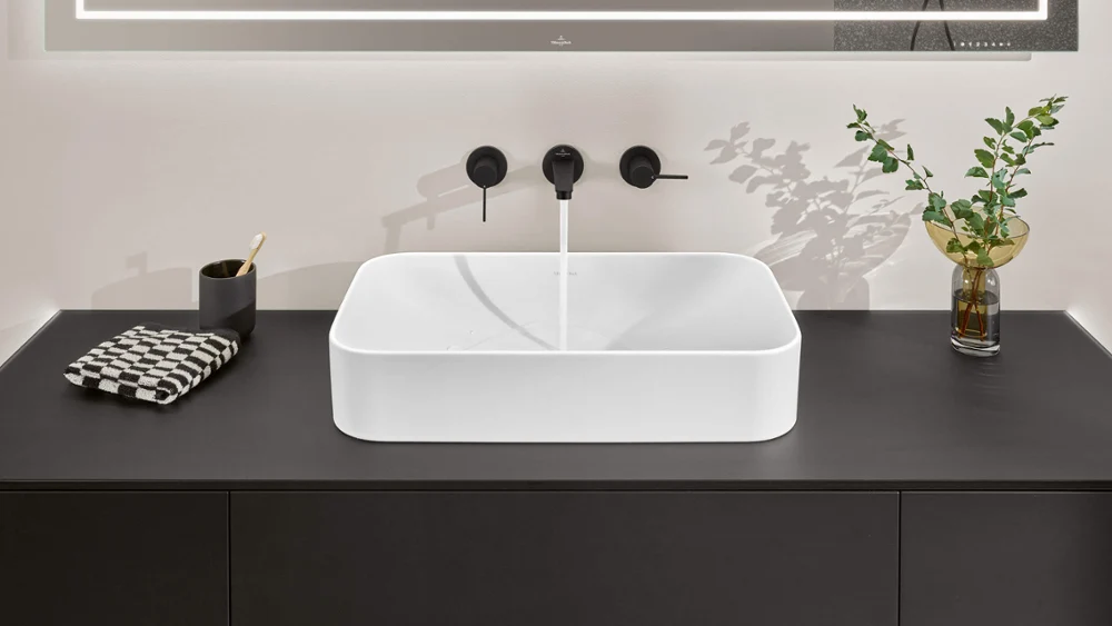 Lavabo de baño moderno con diseño cuadrado sobre encimera negra, grifo cromado y planta decorativa
