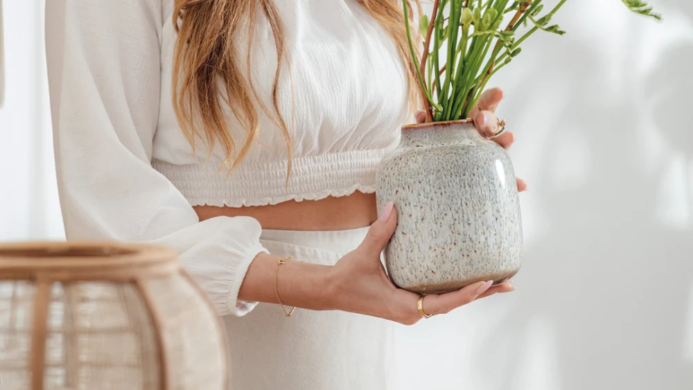 Lave Home Vase as an ideal gift A woman in white clothing hands over a Lave ceramic vase with green and yellow flowers, which stands on a wooden table.