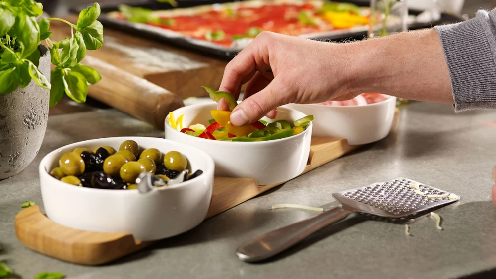 Er staan verschillende kleine witte schaaltjes met olijven en groenten op een aanrecht, met een Kensington Fromage kaasrasp ervoor.