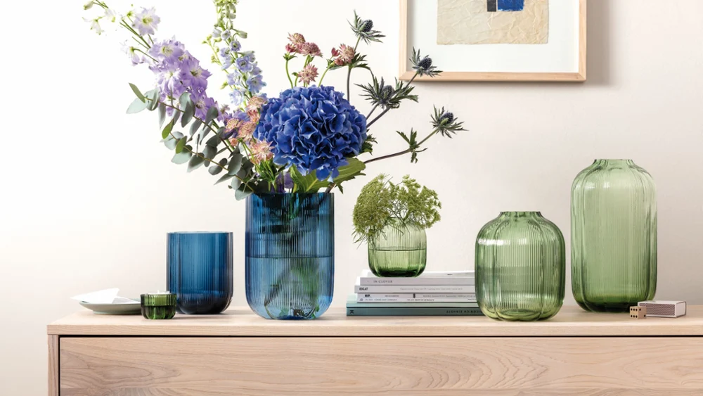 Green and blue glass vases in various shapes some with flowers in them standing on wood furniture with a picture on the wall behind them