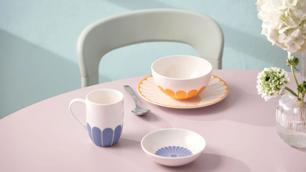 A pastel-coloured table setting with a cup, a bowl and a small plate with blue and orange patterns. A pastel-coloured table setting with a cup, a bowl and a small plate with blue and orange patterns. Between them is a fork, with a bouquet of white flowers in the background.