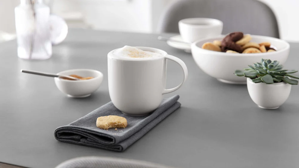 Een kopje cappuccino op een grijs servet met een koekje, schalen met hapjes en een vetplant op een moderne eettafel.