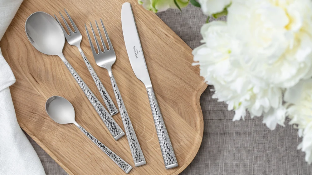 Bovenaanzicht van Blacksmith bestek – soeplepel, tafelvork, tafelmes, koffielepel en dessertvork – netjes gerangschikt op een houten plank. Omlijst door delicate witte bloemdecoratie. Een strakke, natuurlijke en elegante esthetiek.