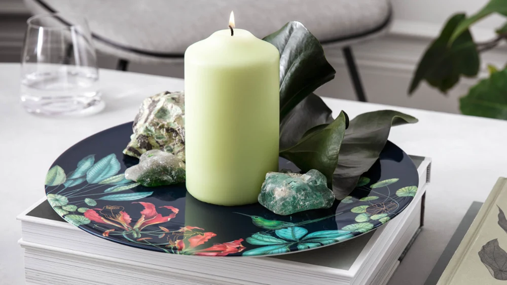 Een stilleven met een brandende kaars, kristal en gebladerte op een decoratief Avarua bord, met boeken en een pen op een moderne tafel.