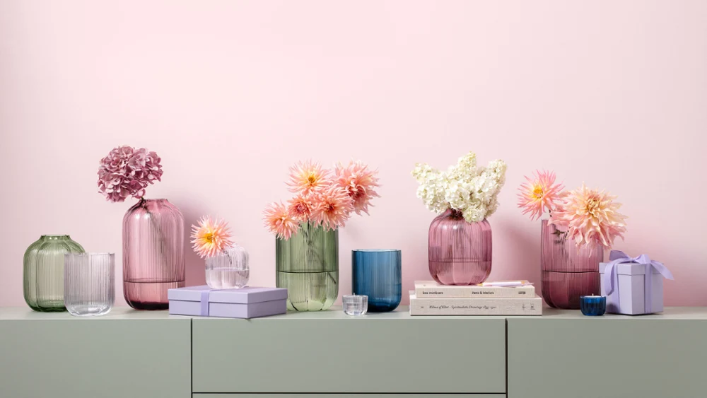 A series of pastel-coloured vases from the Fleur collection in various shapes and sizes with pink, white and peach-coloured flowers, standing on a shelf with stacked books and small boxes.
