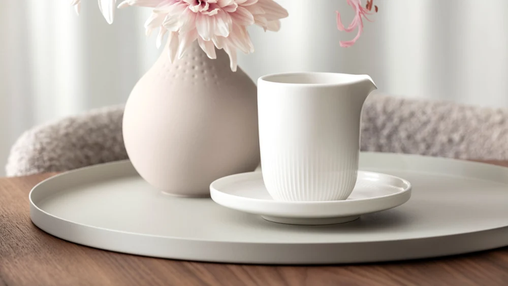 Een witte keramische kan op een schotel staat op een rond dienblad naast een vaas met roze bloemen op een houten tafel.