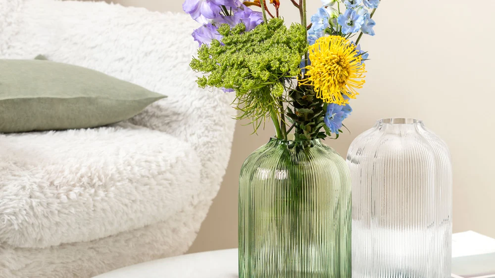 A Villeroy & Boch Fleur vase with colourful flowers and a green vase on a round marble table A round coffee table with a Villeroy & Boch Fleur vase holding blue and green flowers. In the background, an open magazine lies on a white, fluffy chair.
