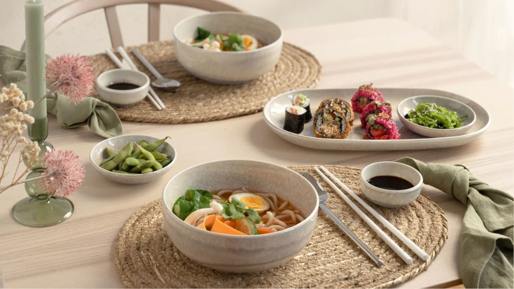 Una mesa puesta para dos personas con piezas Perlemor Una mesa puesta para dos personas con cuencos con ramen, rollitos de sushi, edamame, palillos y salsas para mojar de la colección Perlemor sobre manteles individuales tejidos.