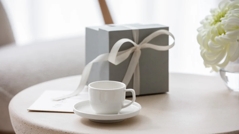 Afina espresso cup and saucer with a gift box A white Afina cup on a saucer, a gift box with a bow and a bouquet of white flowers on a round table.
