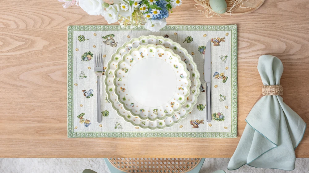 Table setting with stacked Easter Delight plates on a placemat, napkin with ring, and flowers, viewed from above.