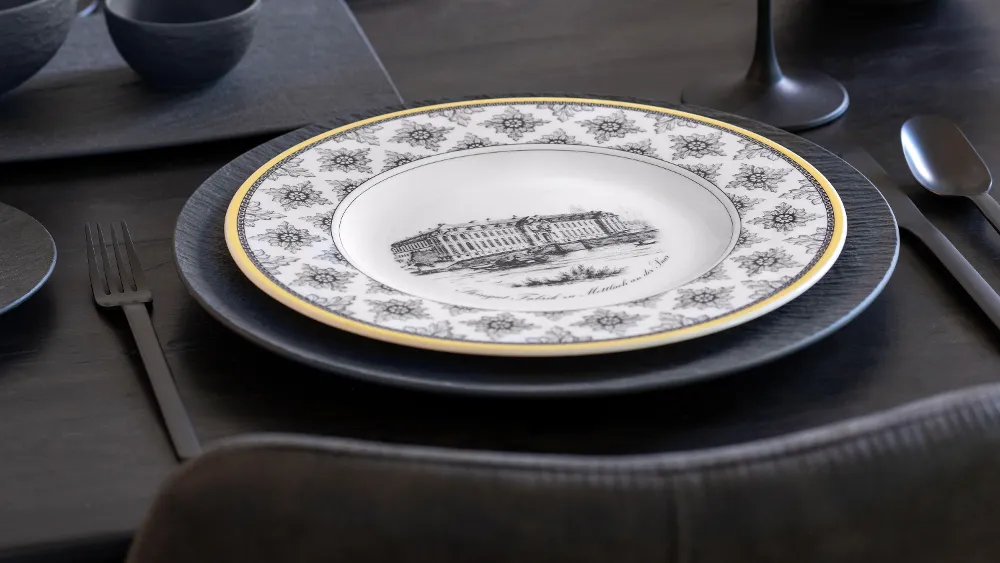 A patterned Audun porcelain plate with a building illustration on a black Manufacture dinner plate, surrounded by black flatware on a dark table setting.