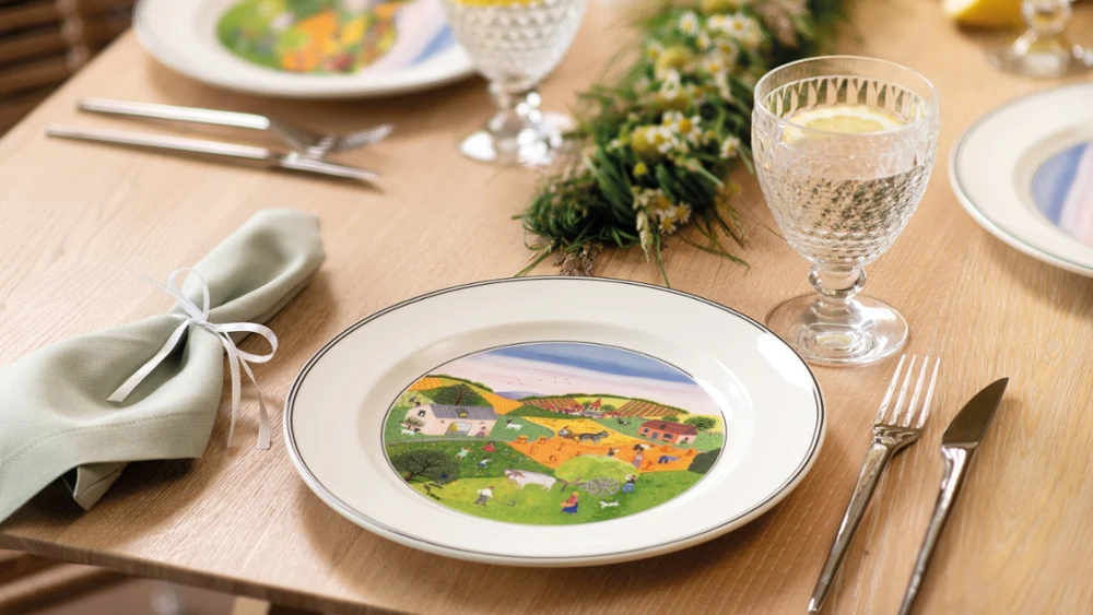 A dining table set with elegant plates featuring colorful countryside scenes. Crystal glasses with lemon slices and a green floral centerpiece add a fresh touch.