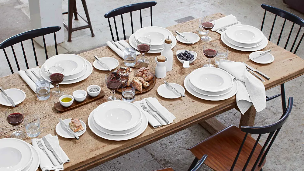 A wooden dining table setting for six people with white crockery, red wine glasses, napkins, bread, grapes, butter, salt, pepper, oil and water glasses. Plates and cutlery are arranged at each place setting.