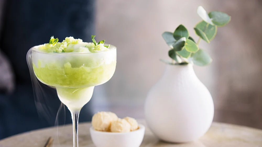 Een gekoelde cocktail, gegarneerd met limoenschil, geserveerd in een Purismo cocktailglas op een marmeren tafel, vergezeld van een kleine schaal met hapjes en een witte vaas met groene planten.