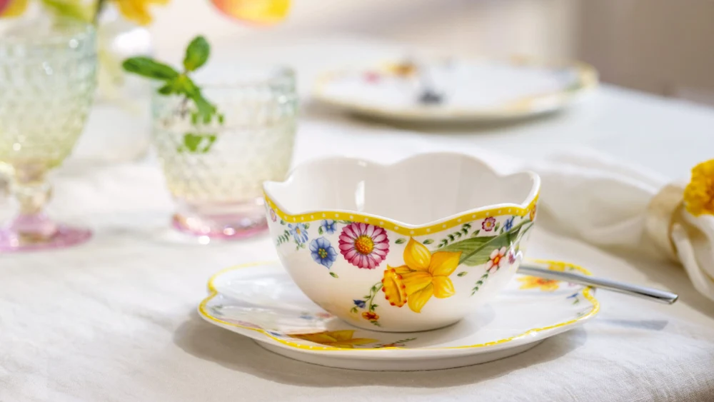 The ‘Spring Awakening’ teacup and saucer with spoon from Villeroy & Boch stand on the table next to crystal glasses and softly blurred flowers in the background.