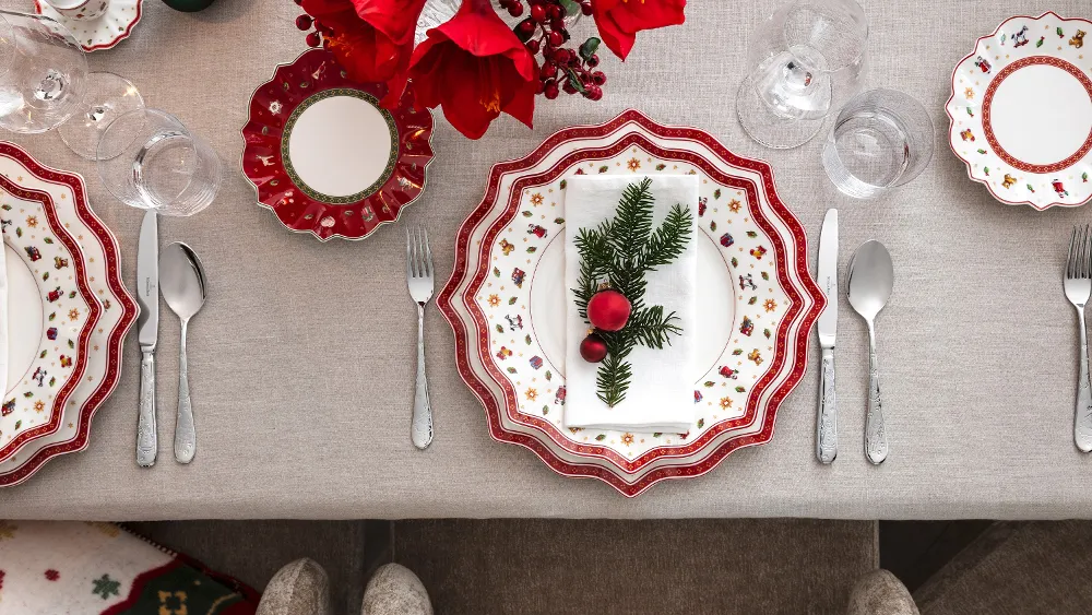 Top view of an elegant festive table with the Villeroy & Boch Toy's Delight collection, complete with red and white plates, silver cutlery and a festive napkin decorated with pine wood and ornaments.