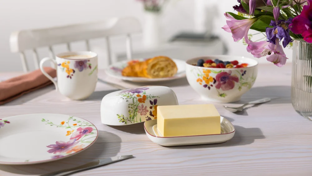 A breakfast table with floral crockery, a butter dish, a cup of coffee, a bowl of fruit, and a plate of pastries.