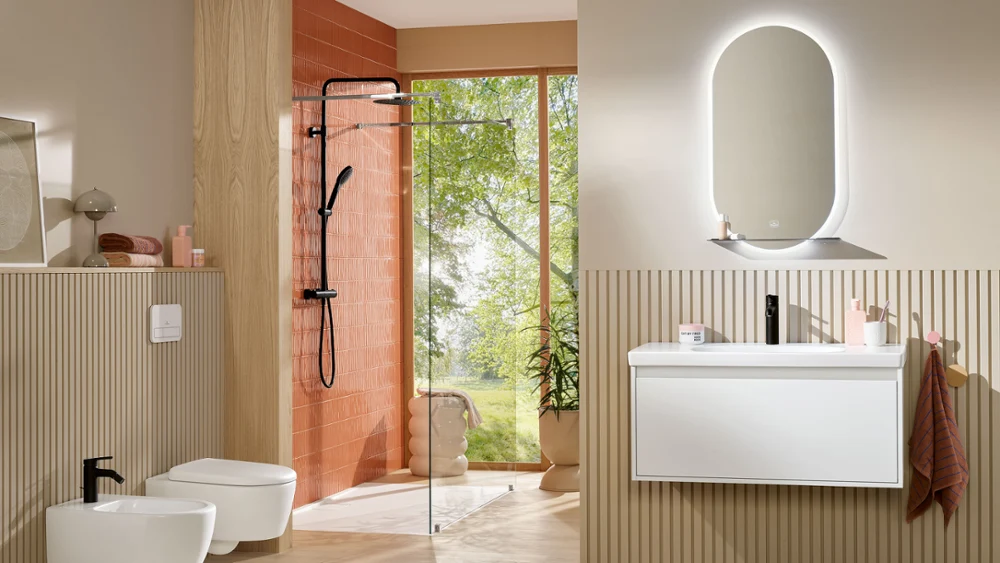 Un elegante baño beige que incluye bidé, inodoro y lavabo Skyla, complementado con una pared de acento de terracota en la zona de ducha. El espejo iluminado More To See Lite añade funcionalidad moderna e iluminación suave y ambiental.