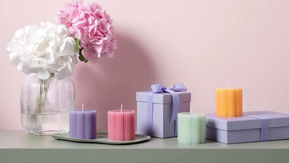 Fleur glass vase with white and pink flowers, pastel-colored Fleur candles in the shape of a cloverleaf, and purple gift boxes on a green surface in front of a pink wall.