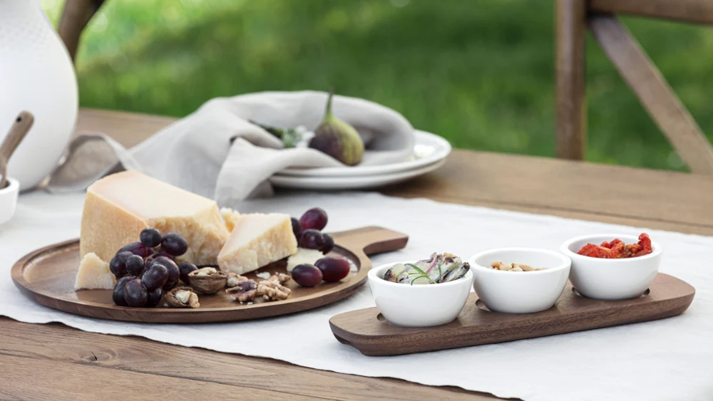 De houten plank uit de Artesano collectie van Villeroy & Boch presenteert kaas, druiven en walnoten, vergezeld van drie kleine schaaltjes met verschillende toppings. Een vaas en gestapelde borden dragen bij aan de esthetiek van de achtergrond.