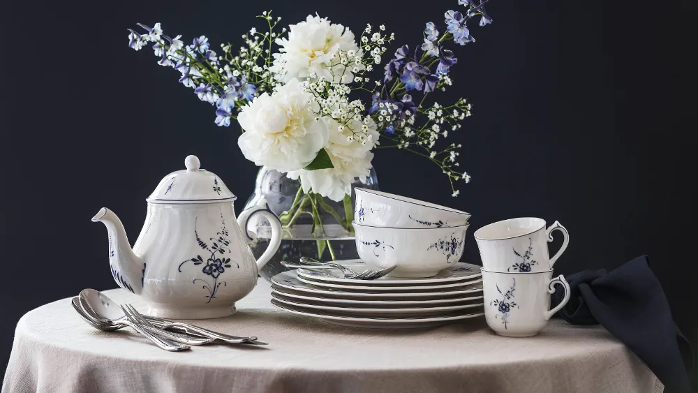 Vieux Luxembourg porcelain on a round table A Vieux Luxembourg porcelain service with a blue and white floral pattern, consisting of a teapot, cups, and plates on a round table, with a vase of white and purple flowers in the background.