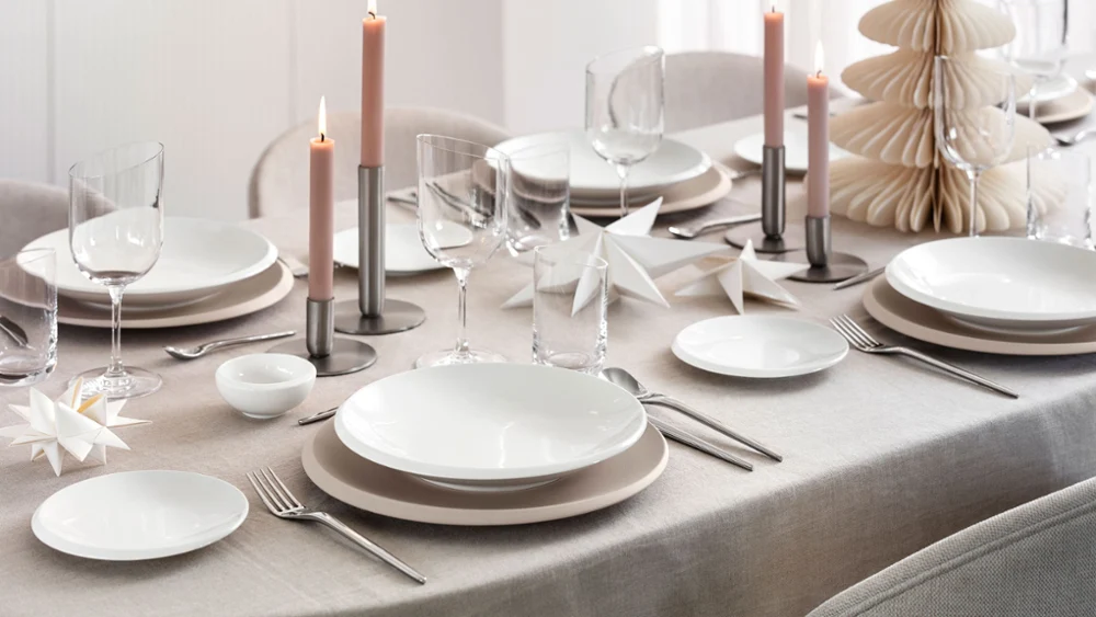 Vista cenital de una mesa festiva moderna con la colección NewMoon Beige de Villeroy & Boch: cuenco plano, platos llanos y de pan, combinados con cubertería NewMoon, copas de vino y vasos. Detalles decorativos navideños suaves, como adornos claros, ramas de pino o velas, aportan un toque estacional refinado a los tonos naturales y formas minimalistas.