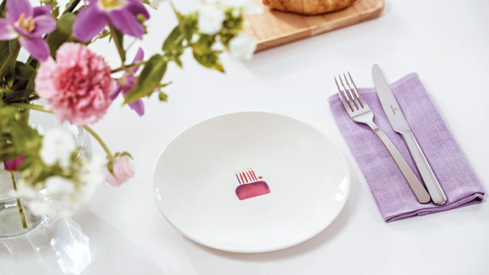 A With Love plate with an illustration of a cake on a table, with a fork and knife on a purple napkin. Next to this is a vase of pink and white flowers and a wooden board with croissants.