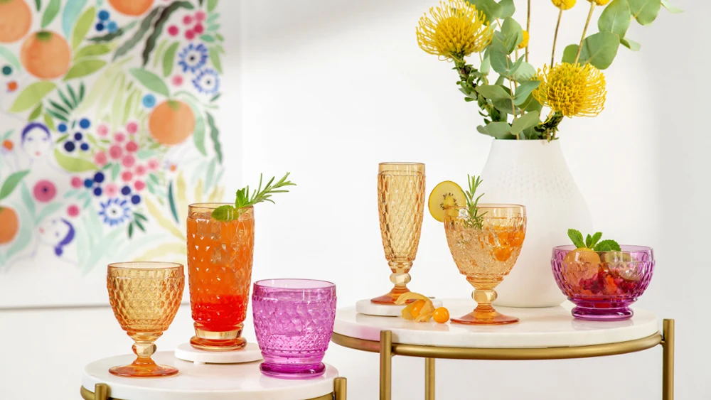 Colourful Boston glasses on tables with various drinks Colourful glasses on tables with various drinks, garnished with herbs and fruit slices. Yellow flowers and green plants in a vase. Wall art with floral and fruit patterns in the background.