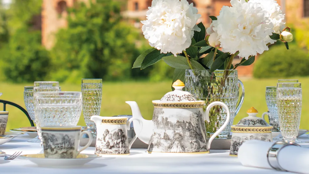 A table set for outdoor tea with ornate teacups and an Audun teapot, crystal glasses, and white peonies in a vase. A historic building is blurred in the background.