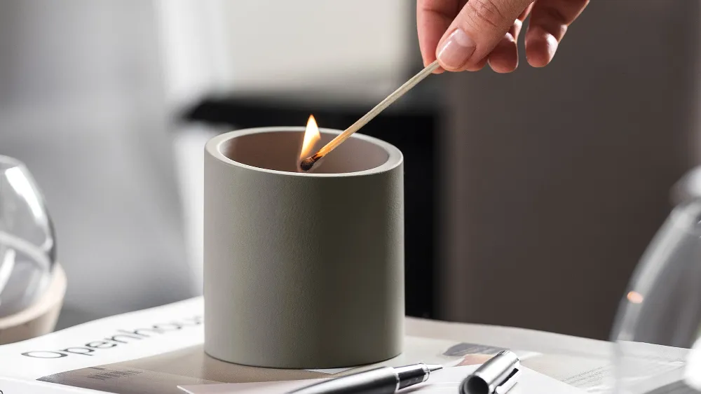 Une main allume une bougie NewMoon de Villeroy & Boch avec une allumette sur une table sur laquelle sont posés du papier et un stylo.