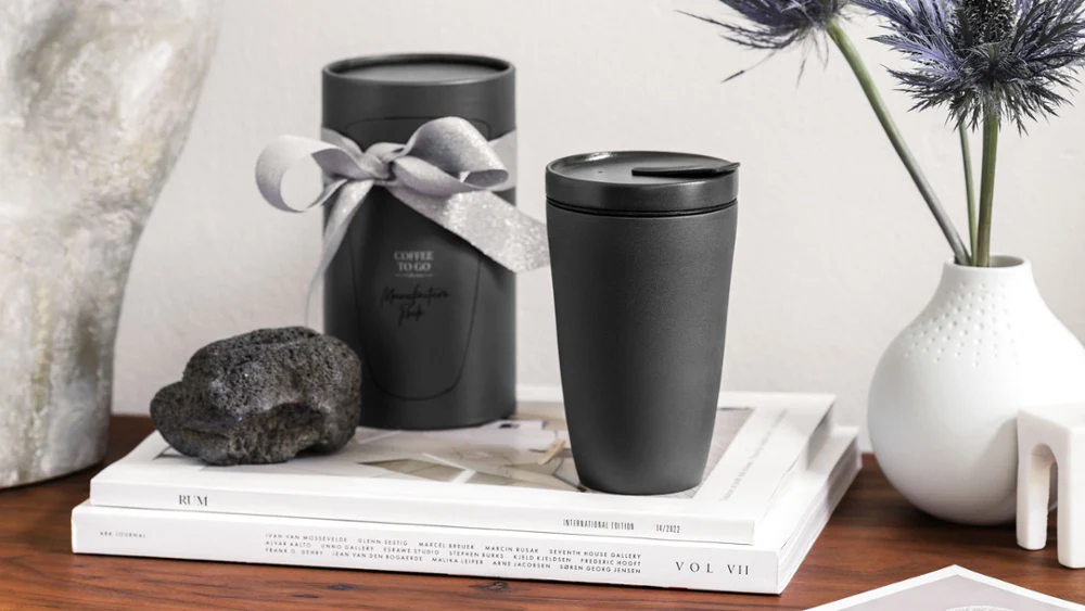 Manufacture To Go coffee mug with decorative objects A stylish workspace with decorative objects, including two black Manufacture To Go coffee mugs, a white vase with blue flowers, stacked books and a stone sculpture on a wooden surface.