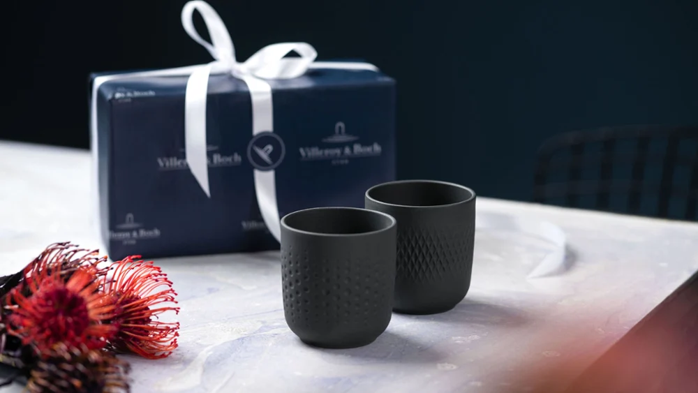 Twee zwarte collierbekers op een tafel met een cadeaudoosje, naast een rode en zwarte bloem.