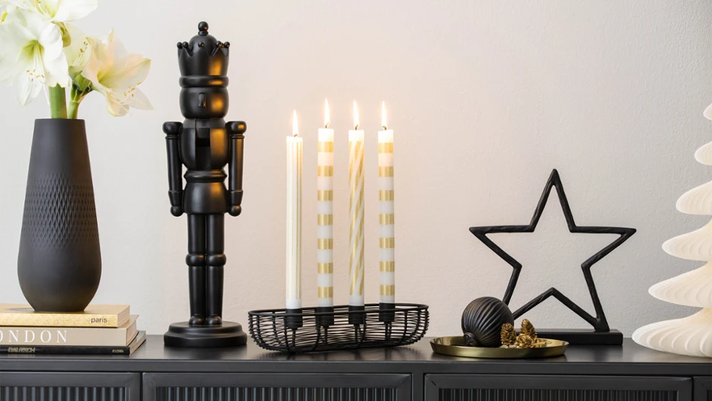 A dark vase, a black nutcracker, four lit candles, star-shaped decorations and small ornaments arranged on a cupboard.