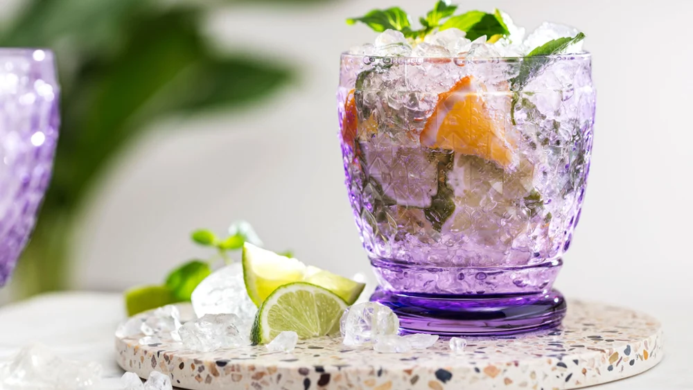 Een verfrissend drankje in een paars Like glas, gegarneerd met limoen en muntblaadjes op een stenen onderzetter.