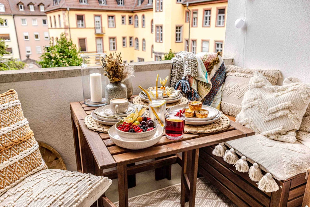Perlemor Sand Breakfast Table on a Cozy Balcony Cozy balcony breakfast setting featuring Perlemor Sand porcelain dinnerware with fresh berries, pastries, coffee, and drinks on a wooden table surrounded by woven textiles and city views.