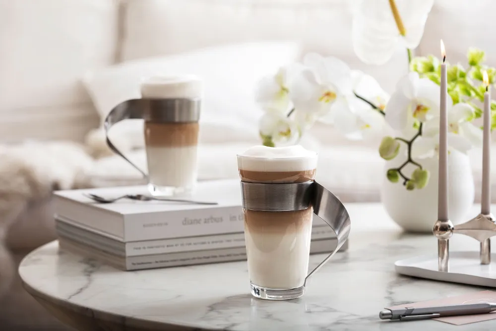 Two coffee drinks in NewWave latte macchiato glasses from Villeroy & Boch with elegant metal handles are placed on a marble table, surrounded by books, orchids and a candlestick.